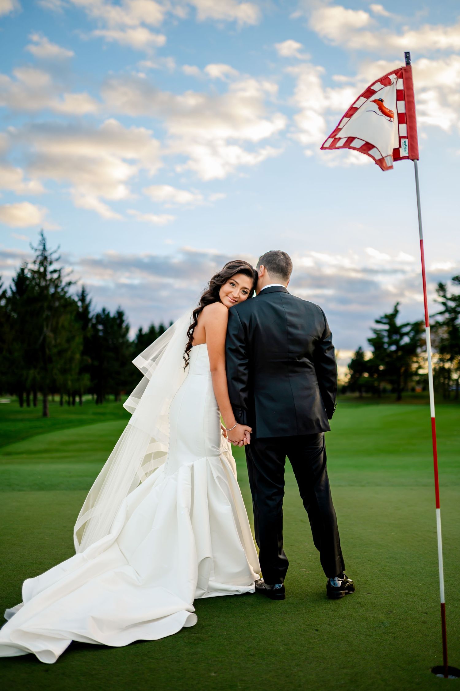 A couple in wedding attire poses playfully near a golf course flag on a sunny day.