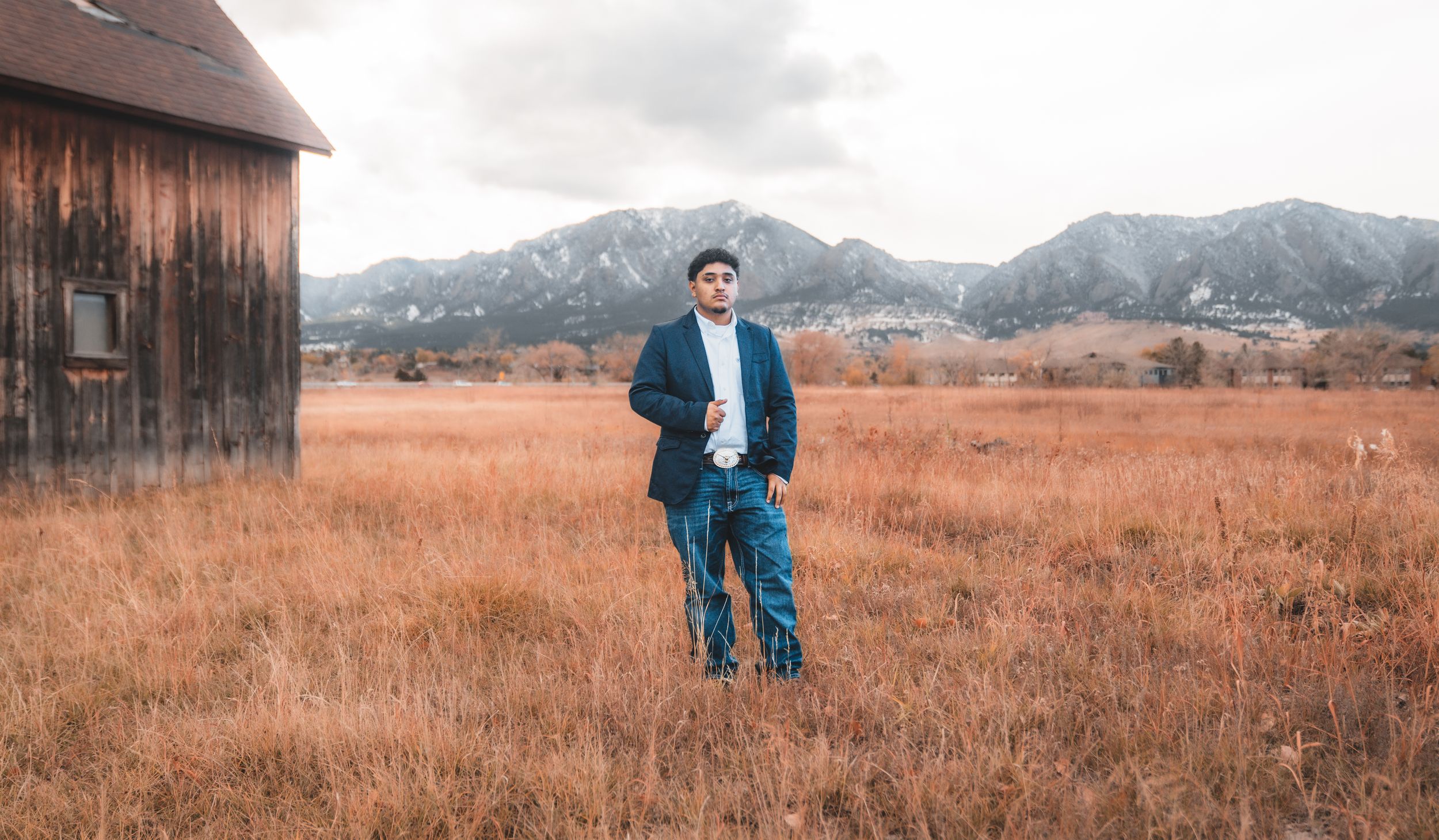 A person stands in a rustic autumn field with mountains and an old barn in the background.
