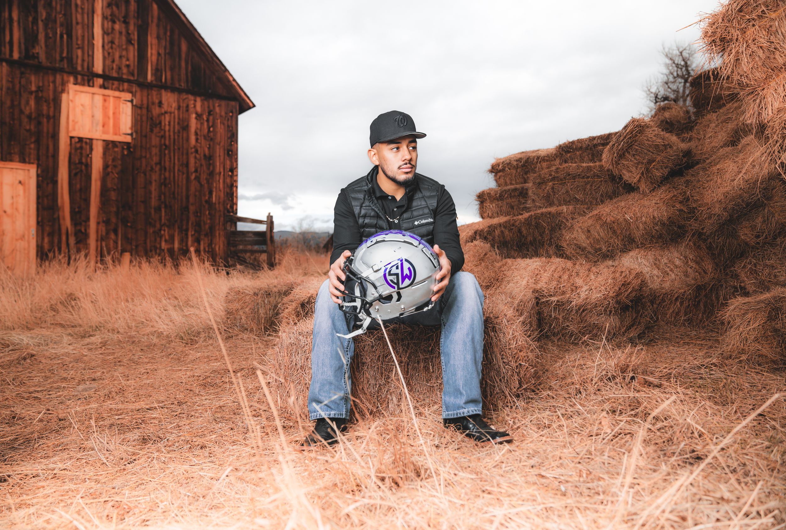 A baseball player in jeans and a cap sits on hay bales near a rustic barn in warm sunset lighting.