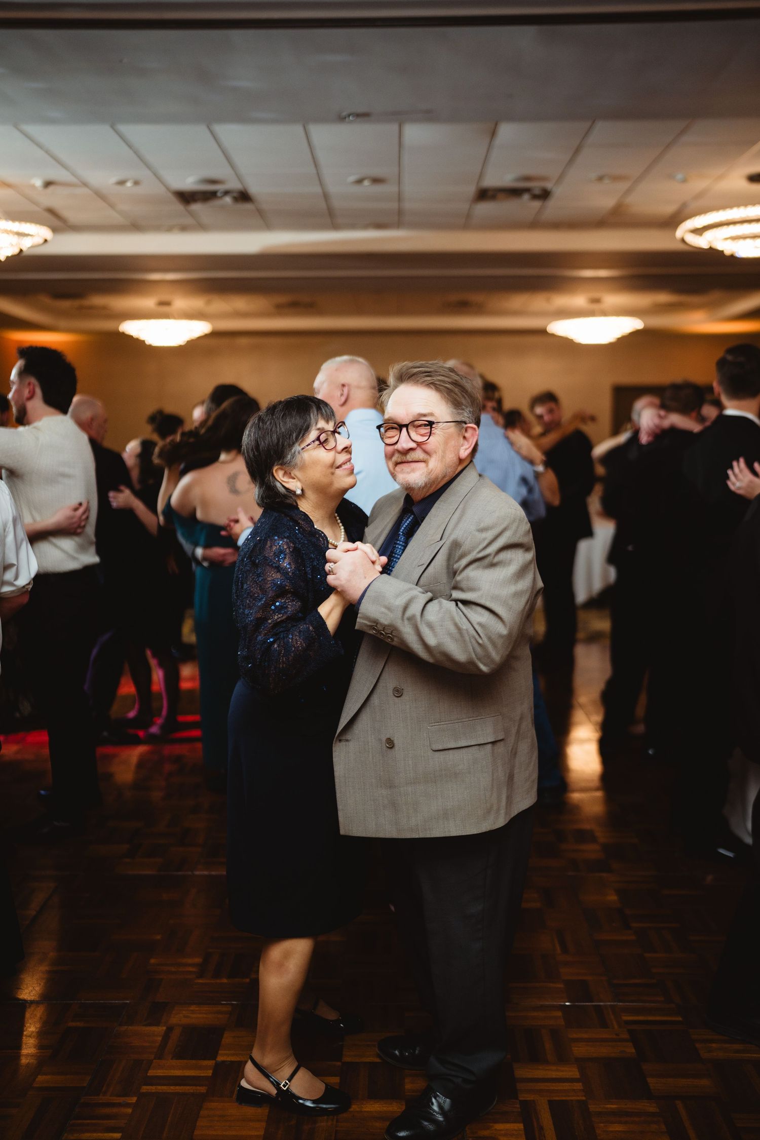 Elderly couples dancing together in a dimly lit ballroom with wood floors and circular ceiling lights.