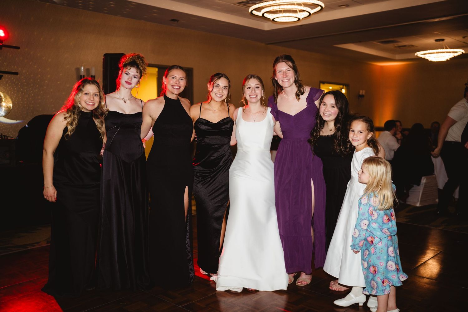 A group of wedding guests dance and celebrate together on a dimly lit dance floor at an evening reception.