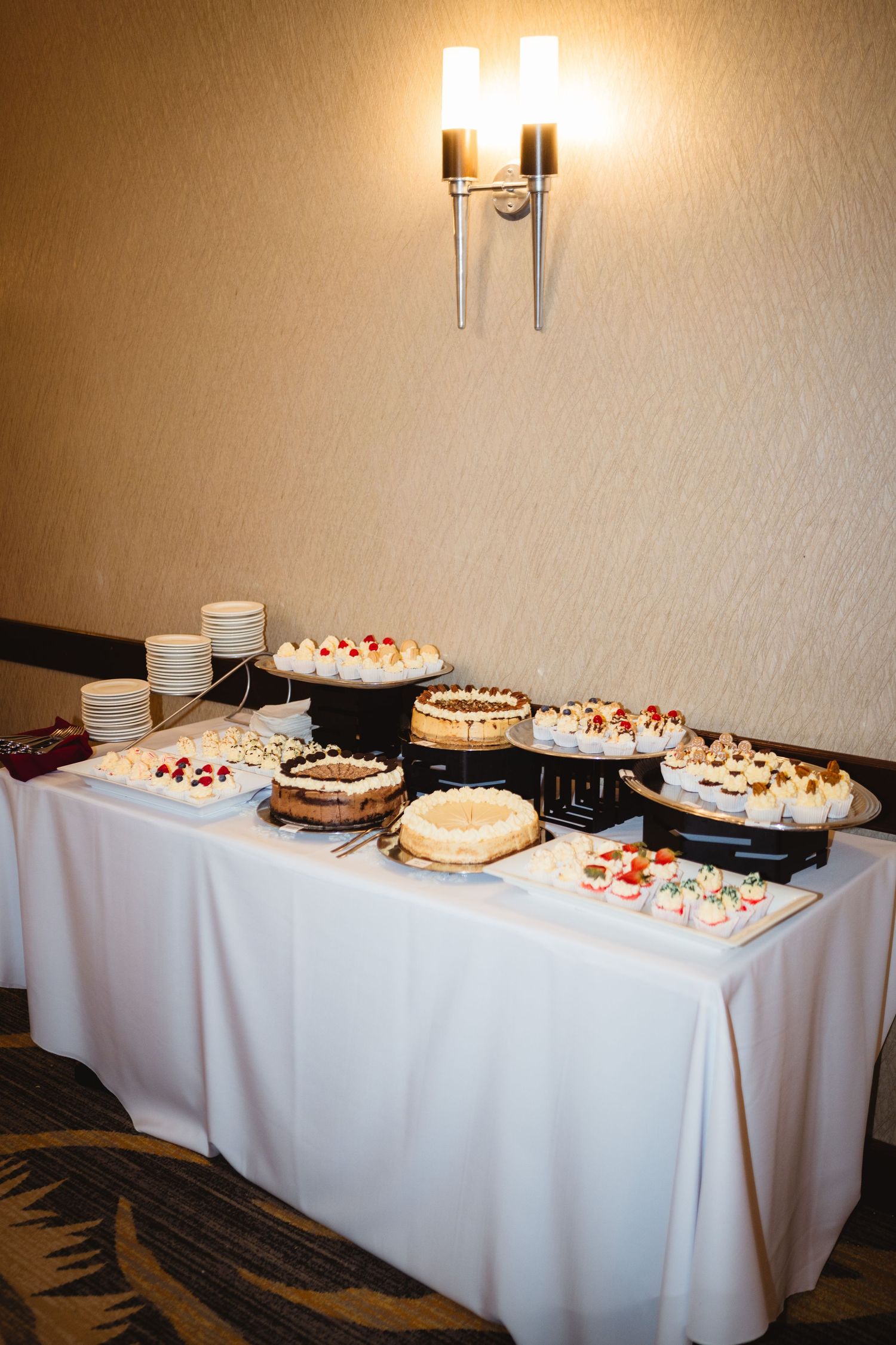 A panoramic view of dessert cakes displayed on a long white-clothed table with warm lighting in an event space.