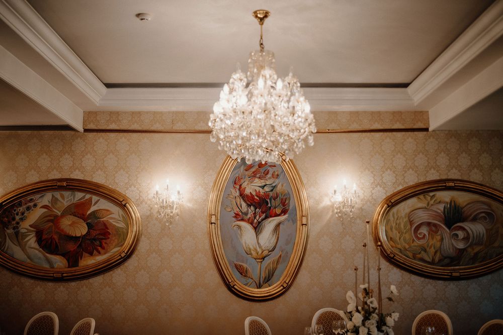 Crystal chandeliers hang elegantly from an ornate ceiling decorated with gold trim and painted medallions.