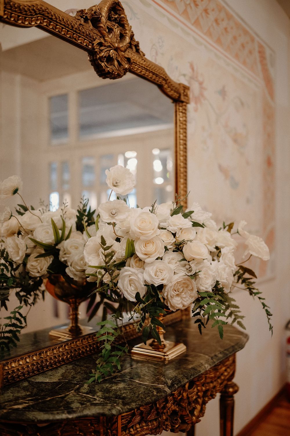 White roses and greenery arranged on an ornate gold-framed mirror create an elegant wedding decor display.