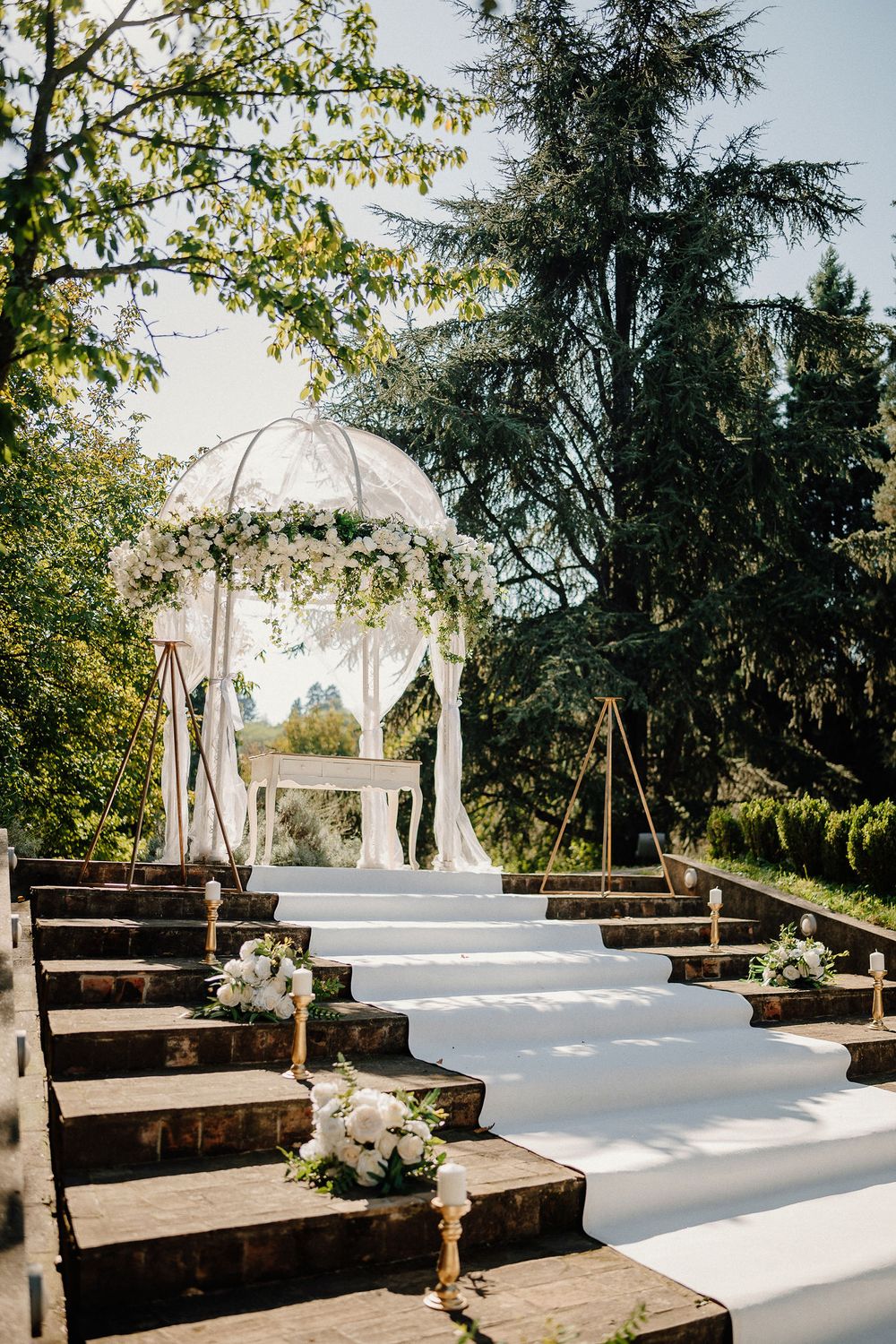 Outdoor wedding ceremony setup with wooden benches and white domed gazebo surrounded by evergreen trees.