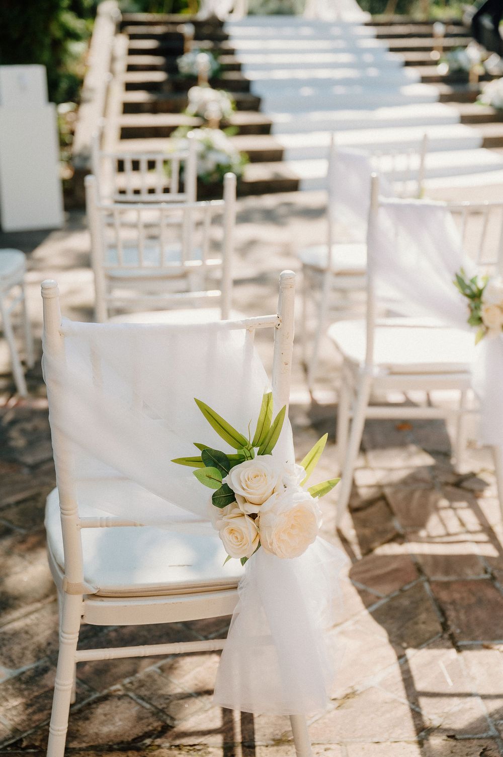 White Chiavari chairs decorated with roses and tulle line up at an outdoor wedding ceremony on a stone patio.