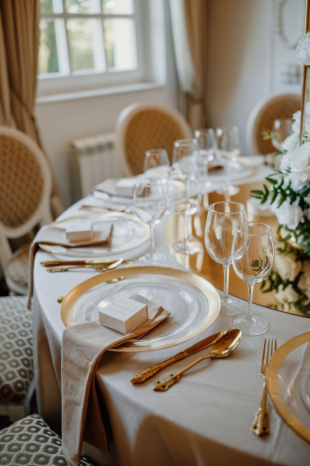 Elegant wedding table settings with gold cutlery, white linens, and crystal wine glasses in natural window light.