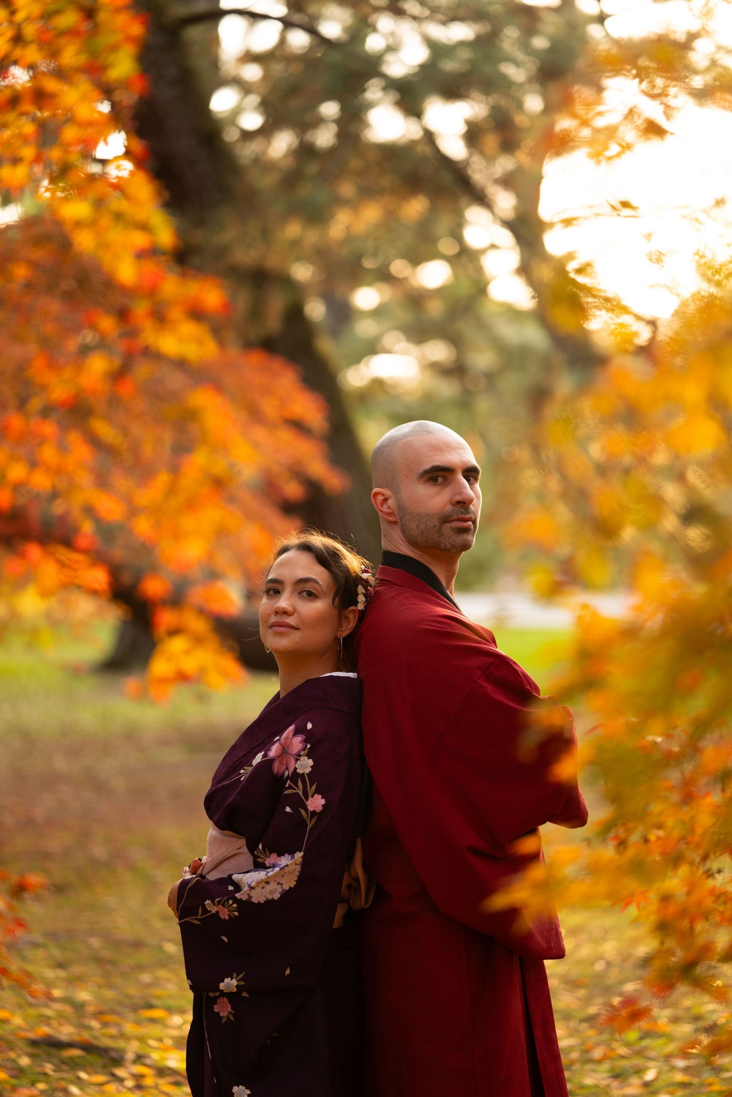 Couple in matching dark and red kimono standing back-to-back among breathtaking golden autumn trees in Japan.