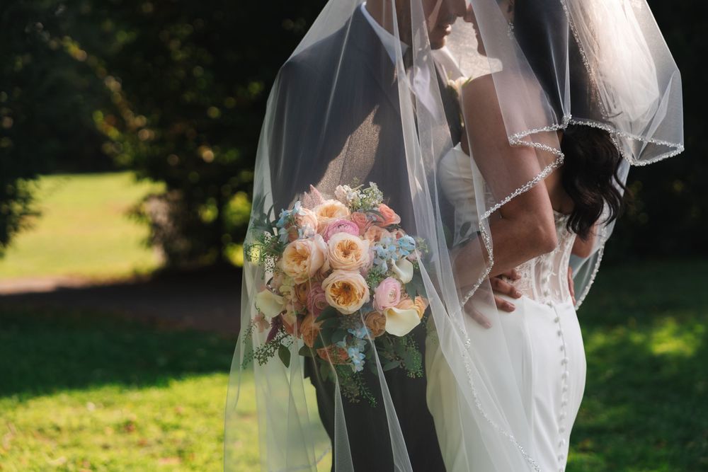 A wedding bouquet with peach and white flowers held by someone in a flowing white wedding dress and veil outdoors.