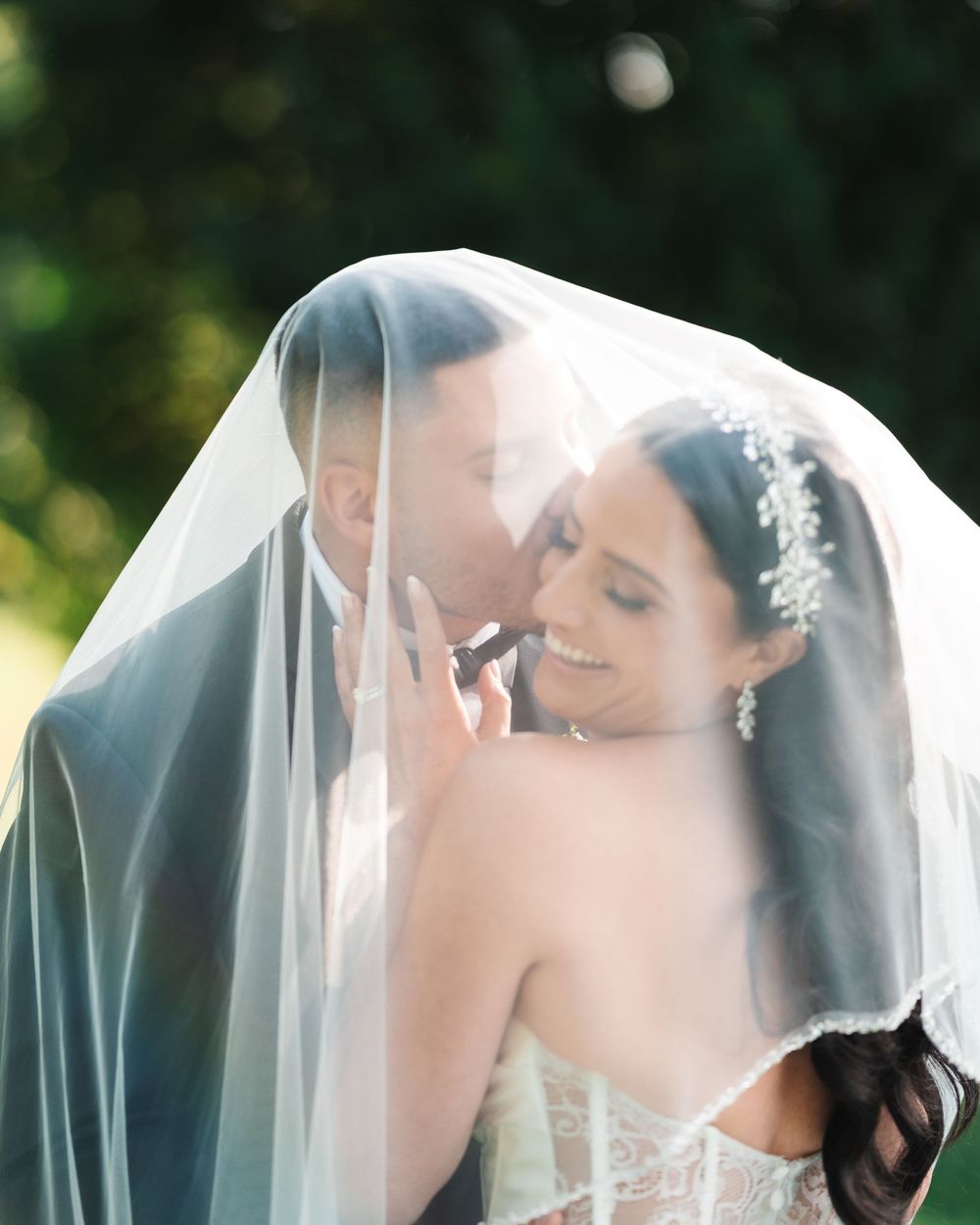 A romantic wedding moment captured through a flowing white veil in an outdoor garden setting.
