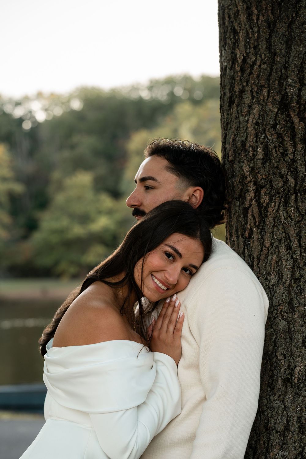 A romantic couple embraces against a tree trunk by a lake at sunset, with the person in white wearing an off-shoulder dress.