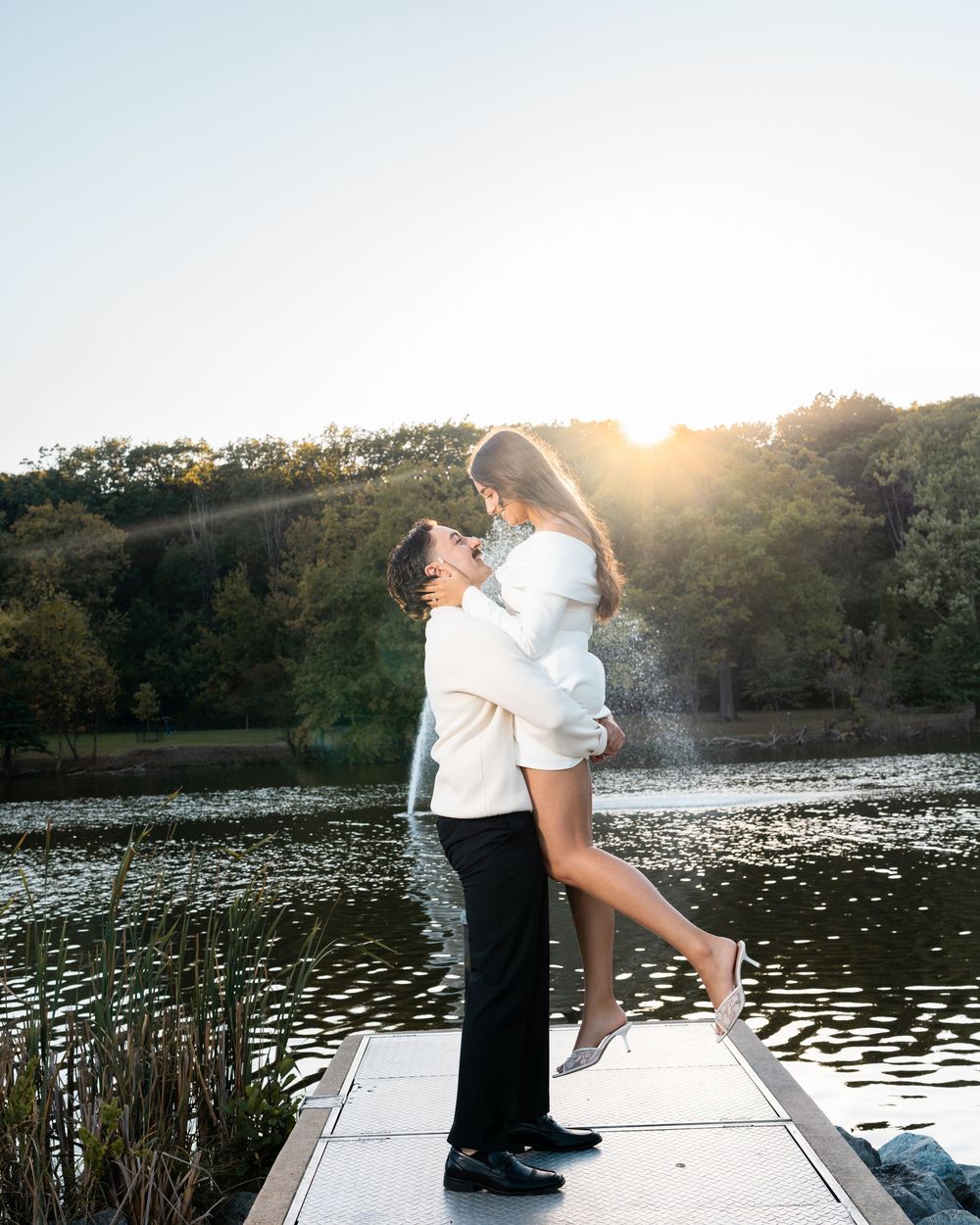 A romantic couple embraces on a wooden dock by a serene lake during golden hour sunset.