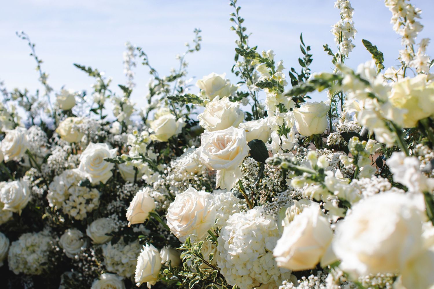 A delicate white floral arch arrangement with roses and greenery creates an elegant wedding ceremony backdrop.