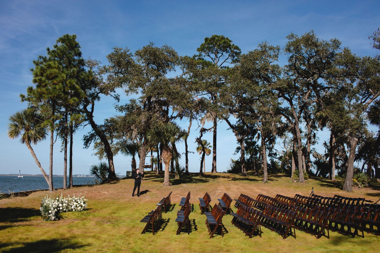 Outdoor wedding ceremony setup with rows of chairs facing the water under tall palm trees and oak trees.