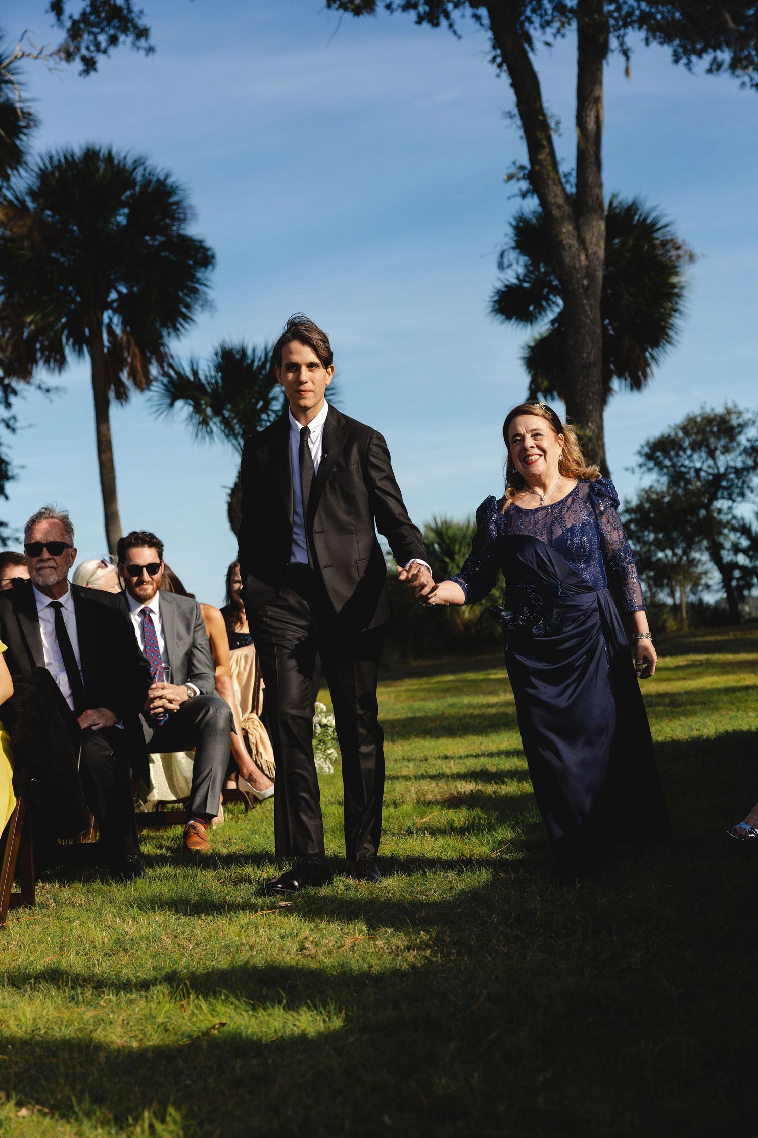 Wedding guests walk across a grassy lawn with palm trees under blue skies.