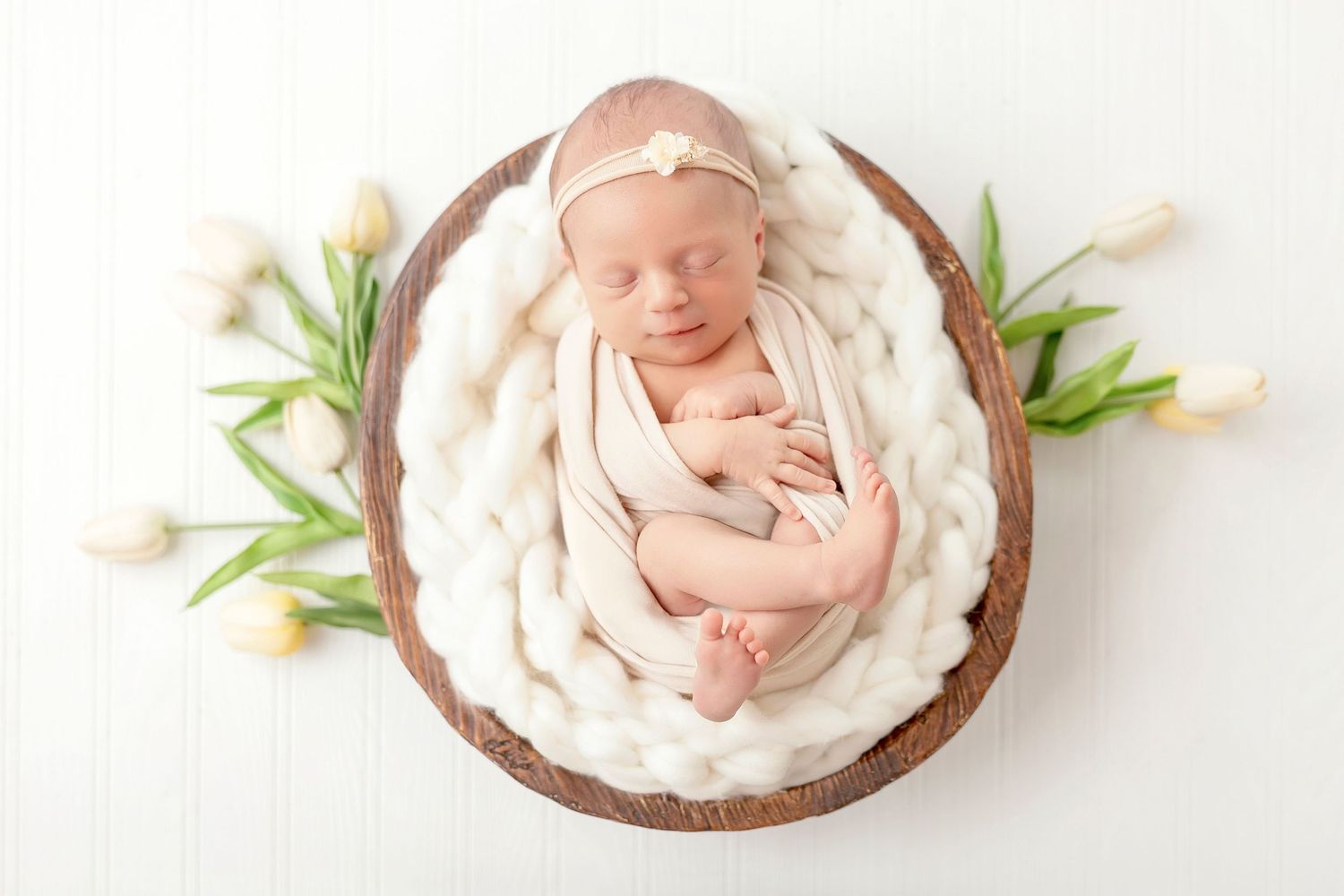 Newborn baby sleeps peacefully in round wooden bowl with white fabric and tulip flowers on white background.