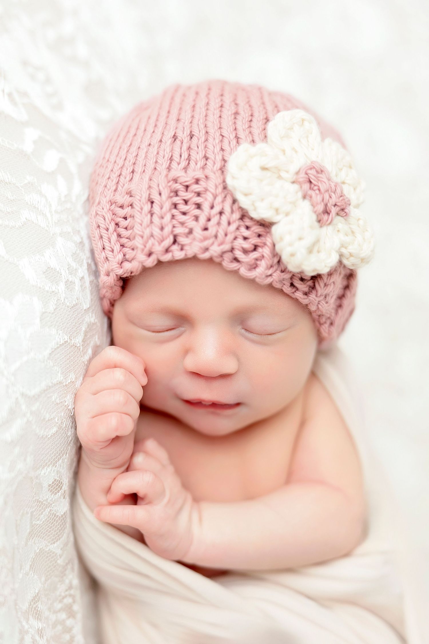 Sleeping newborn wearing soft pink knit beanie with white flower detail wrapped in white blanket on lace background.