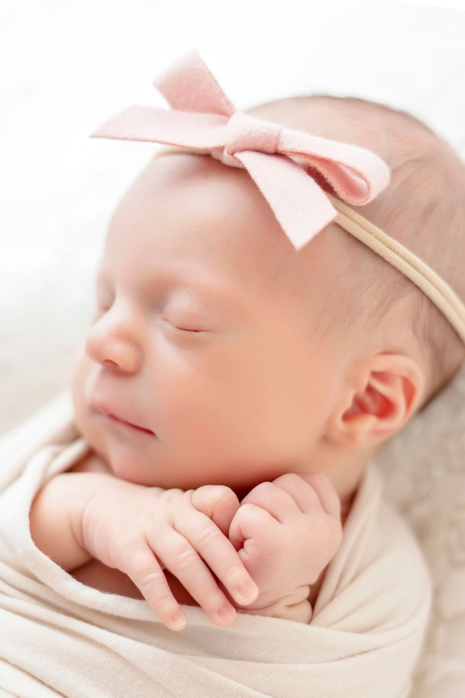 A peaceful sleeping newborn baby wearing a pink bow headband rests on soft white fabric.