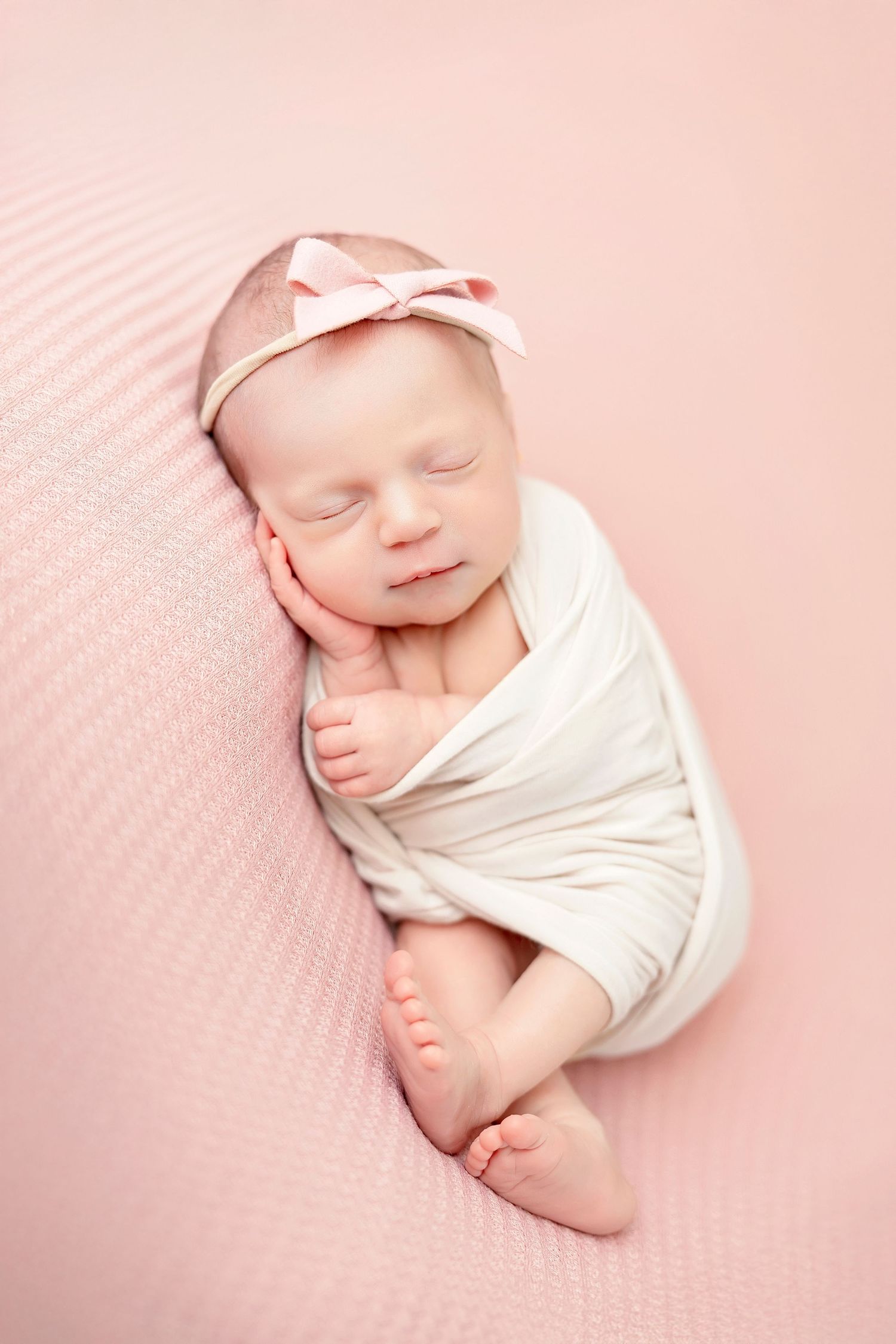Sleeping newborn baby wrapped in white cloth lying on soft pink textured blanket in gentle natural light.