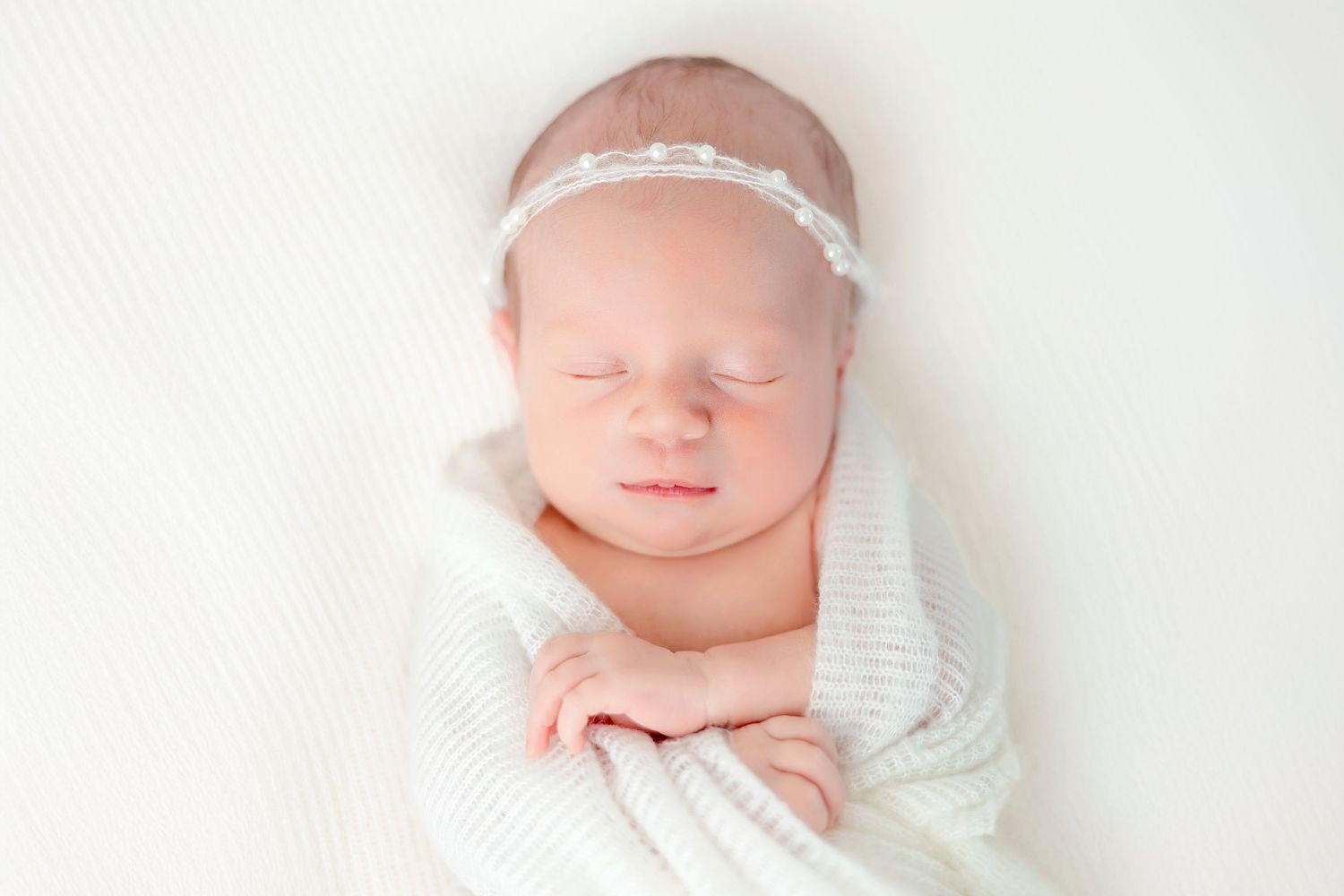 A peaceful sleeping newborn baby wrapped in white swaddling cloth wearing a delicate lace headband.