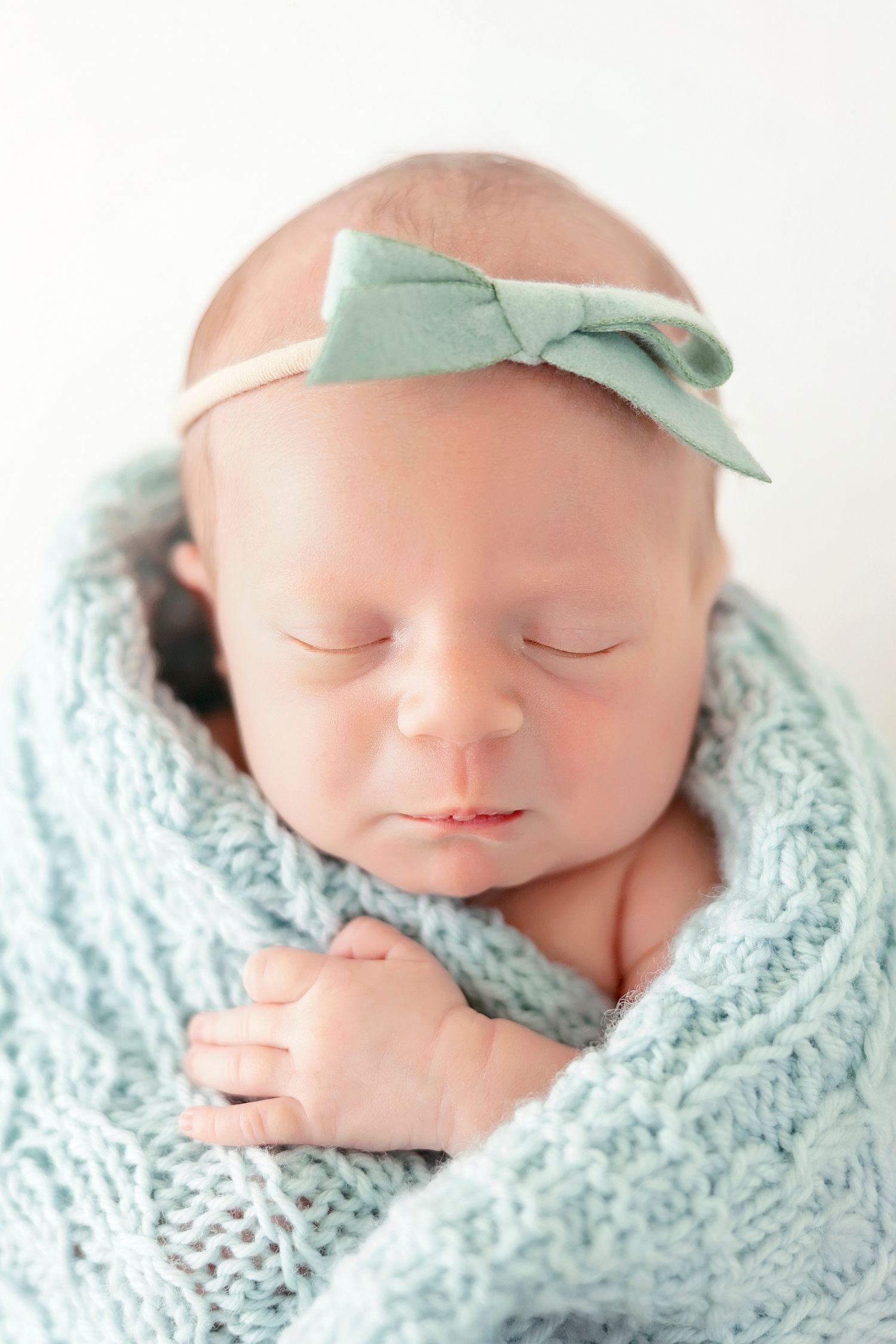 Sleeping newborn wrapped in soft mint green knit blanket wearing matching bow headband in professional portrait series.