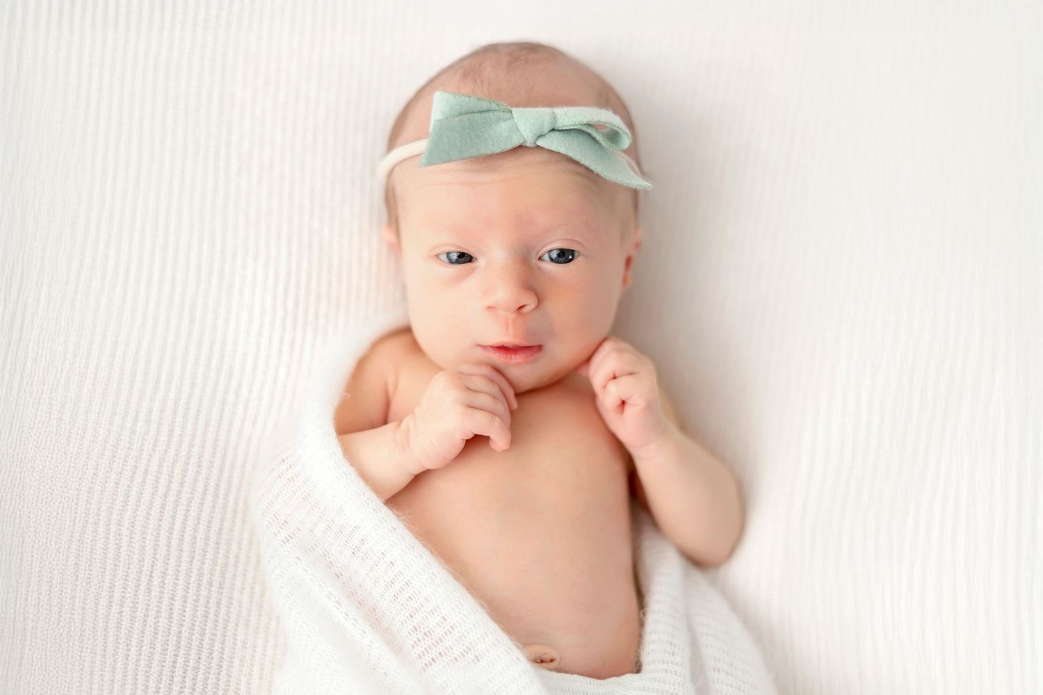 Adorable newborn baby wearing a mint green bow headband lying on white textured fabric.