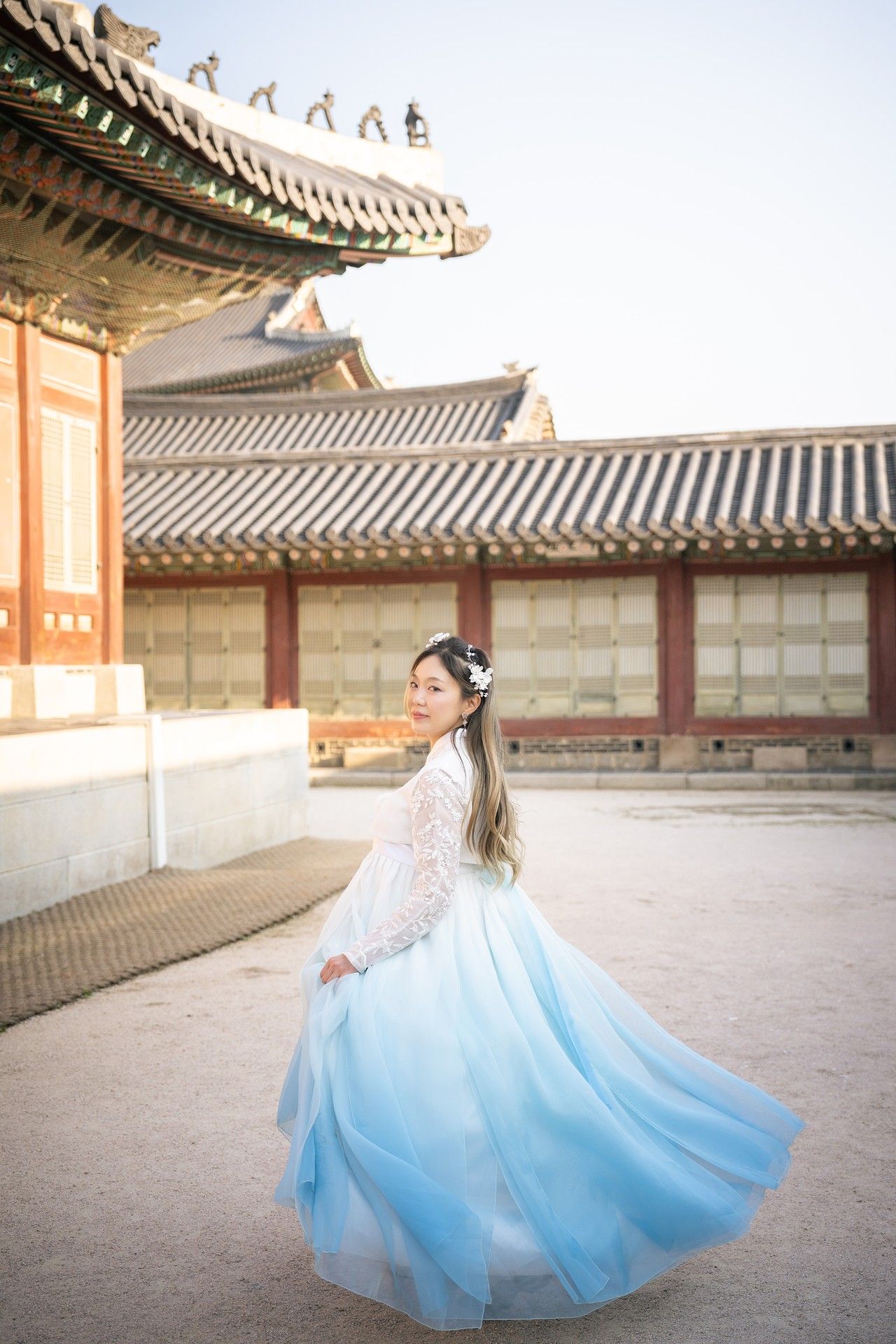 A figure in a flowing white and blue hanbok dress twirls gracefully in a traditional Korean palace courtyard.