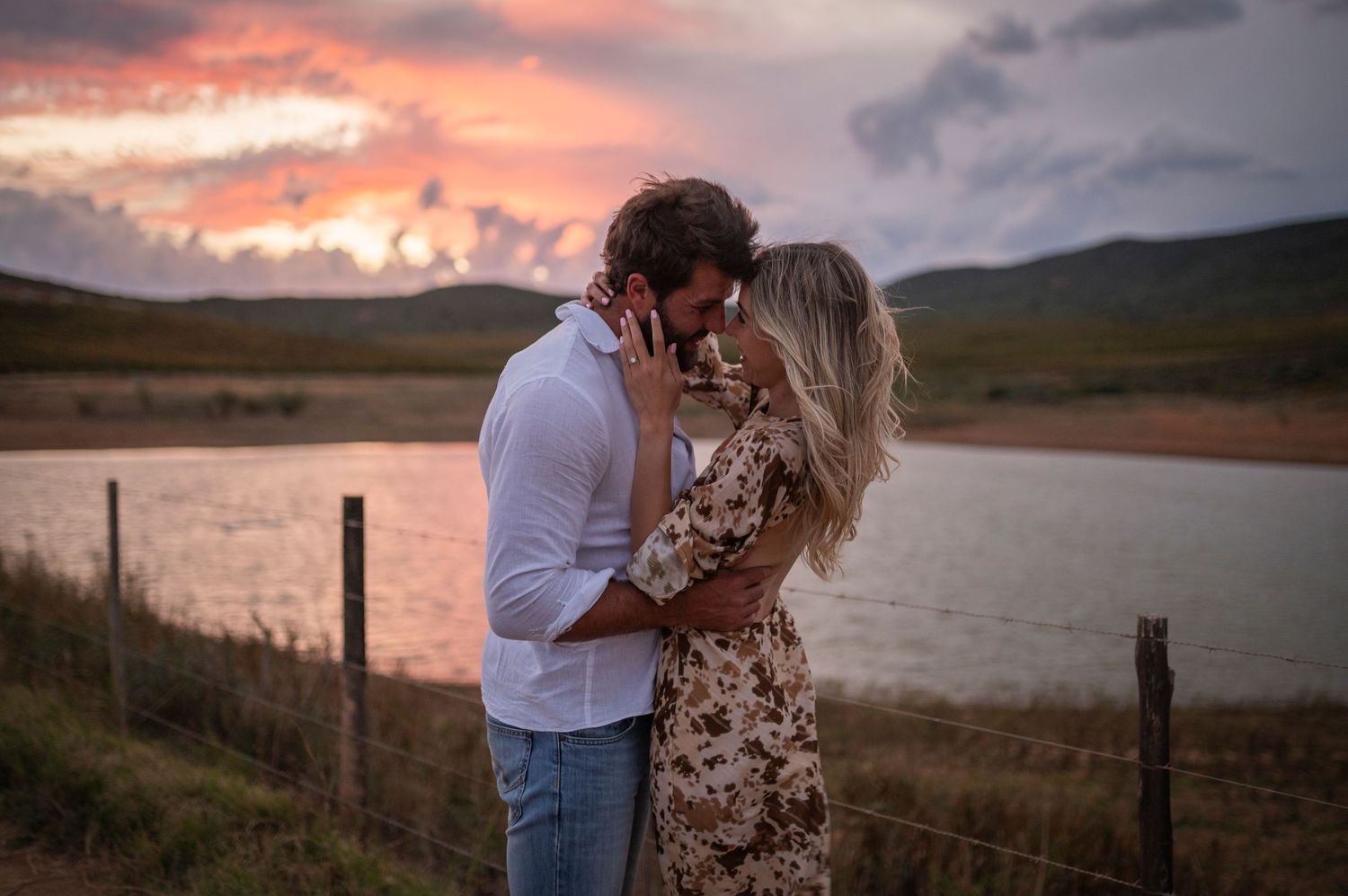 A couple embraces during a romantic sunset photoshoot by a lake with mountains in the background.