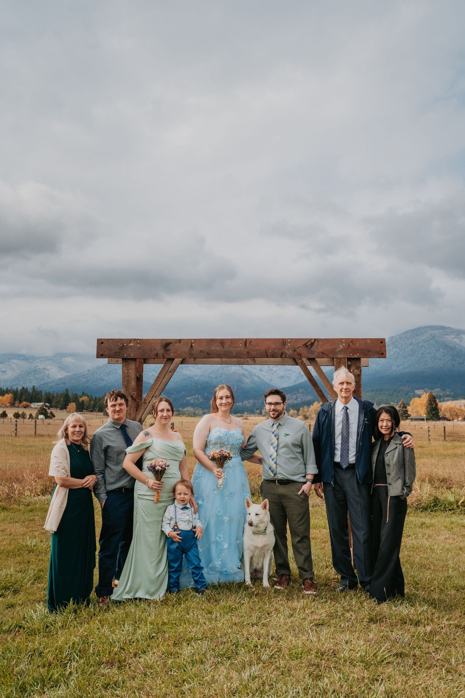 Wedding party stands together under wooden arch with mountain landscape and cloudy sky in background.