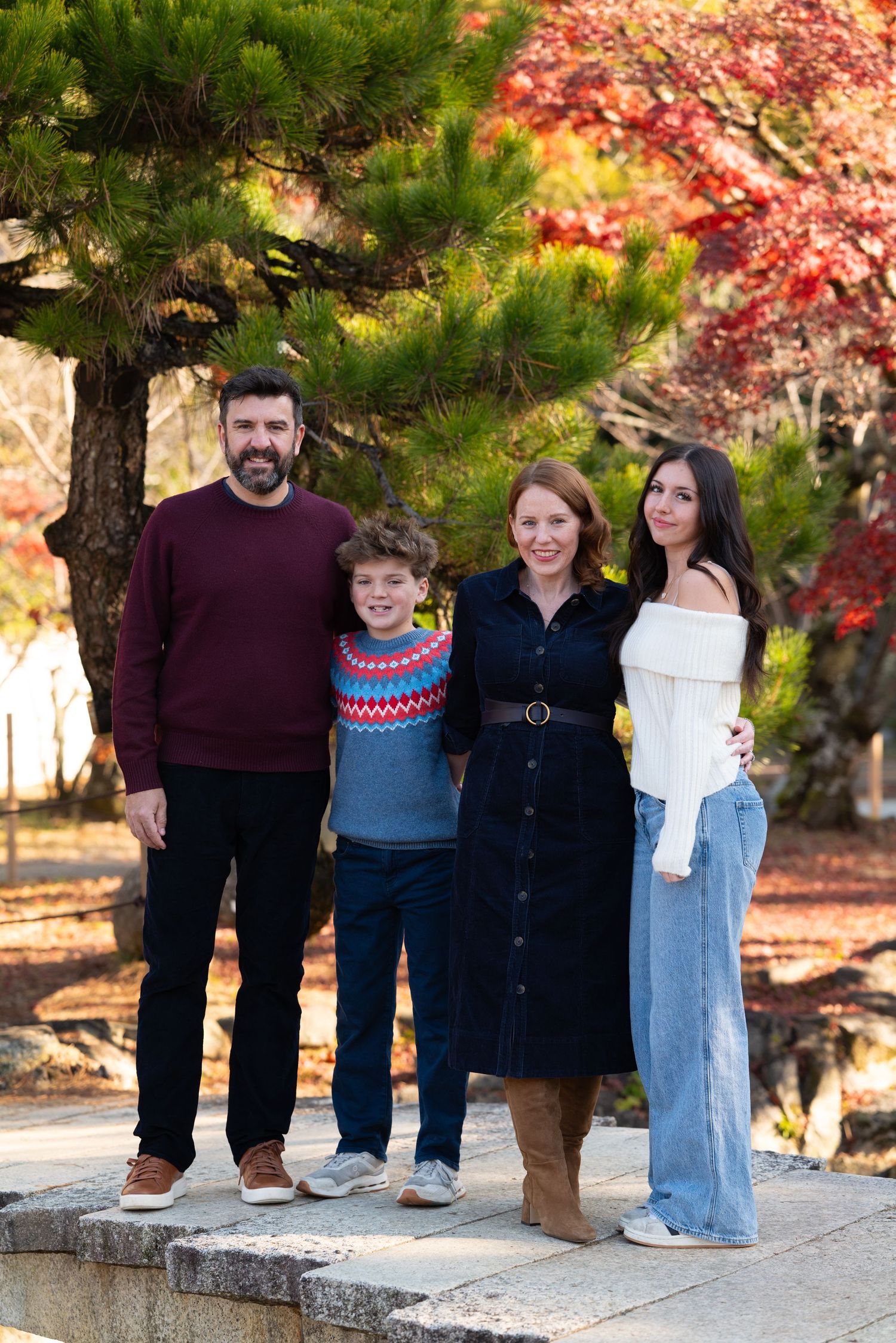 Family of four posing near a pine tree with colorful autumn foliage in Japan.