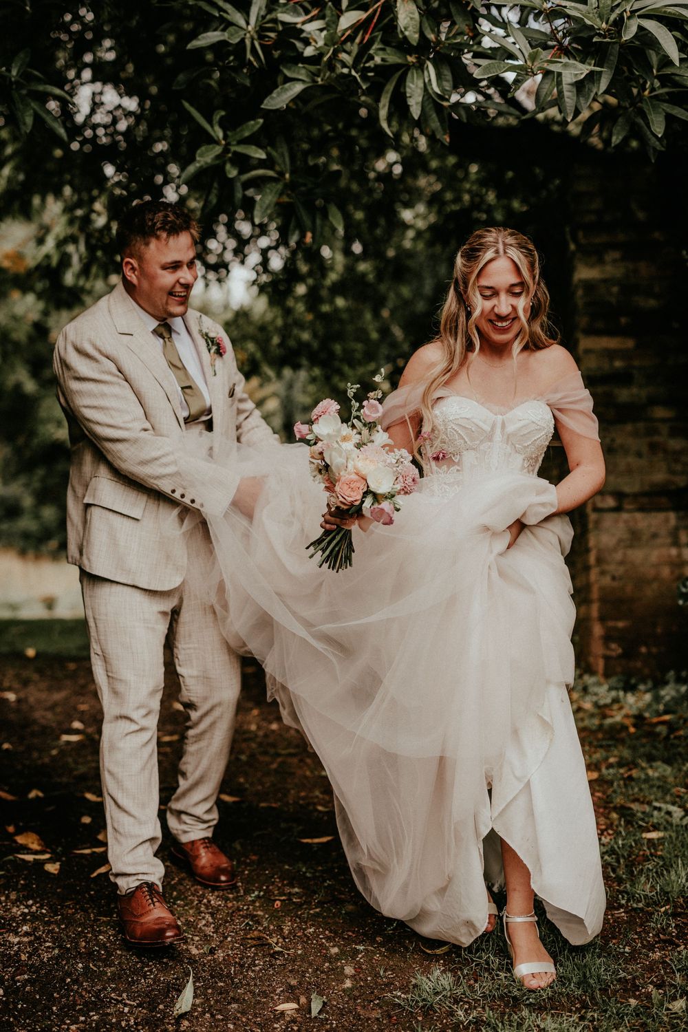 A couple in wedding attire playfully walking together through a garden setting with natural greenery in the background.