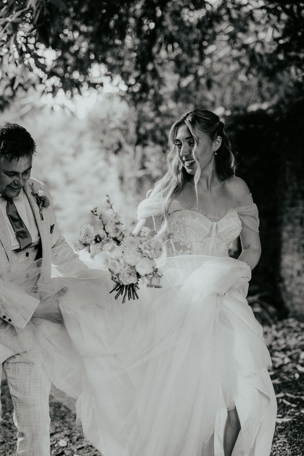 Black and white photo of bridesmaids in flowing white dresses holding bouquets while walking through a garden setting.