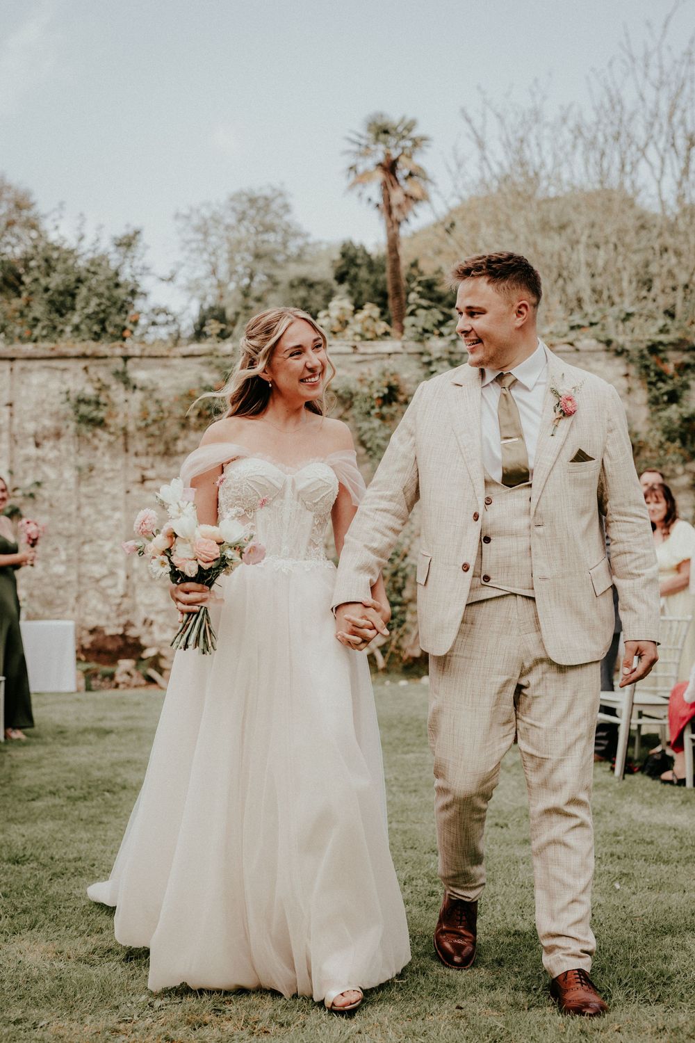 A wedding couple walks and dances together on a grassy lawn in front of a stone wall wearing light colored attire. Cornwall wedding photogra