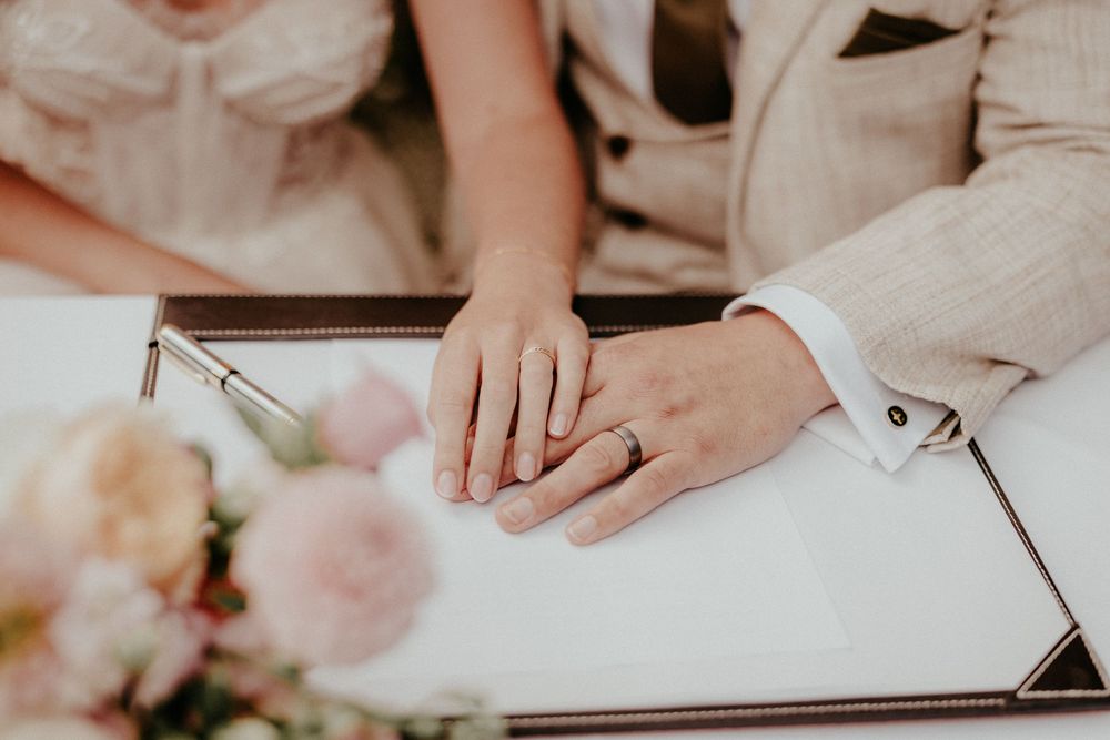 Close-up of hands touching while signing wedding documents with pink flowers in the foreground. Cornwall wedding photographer