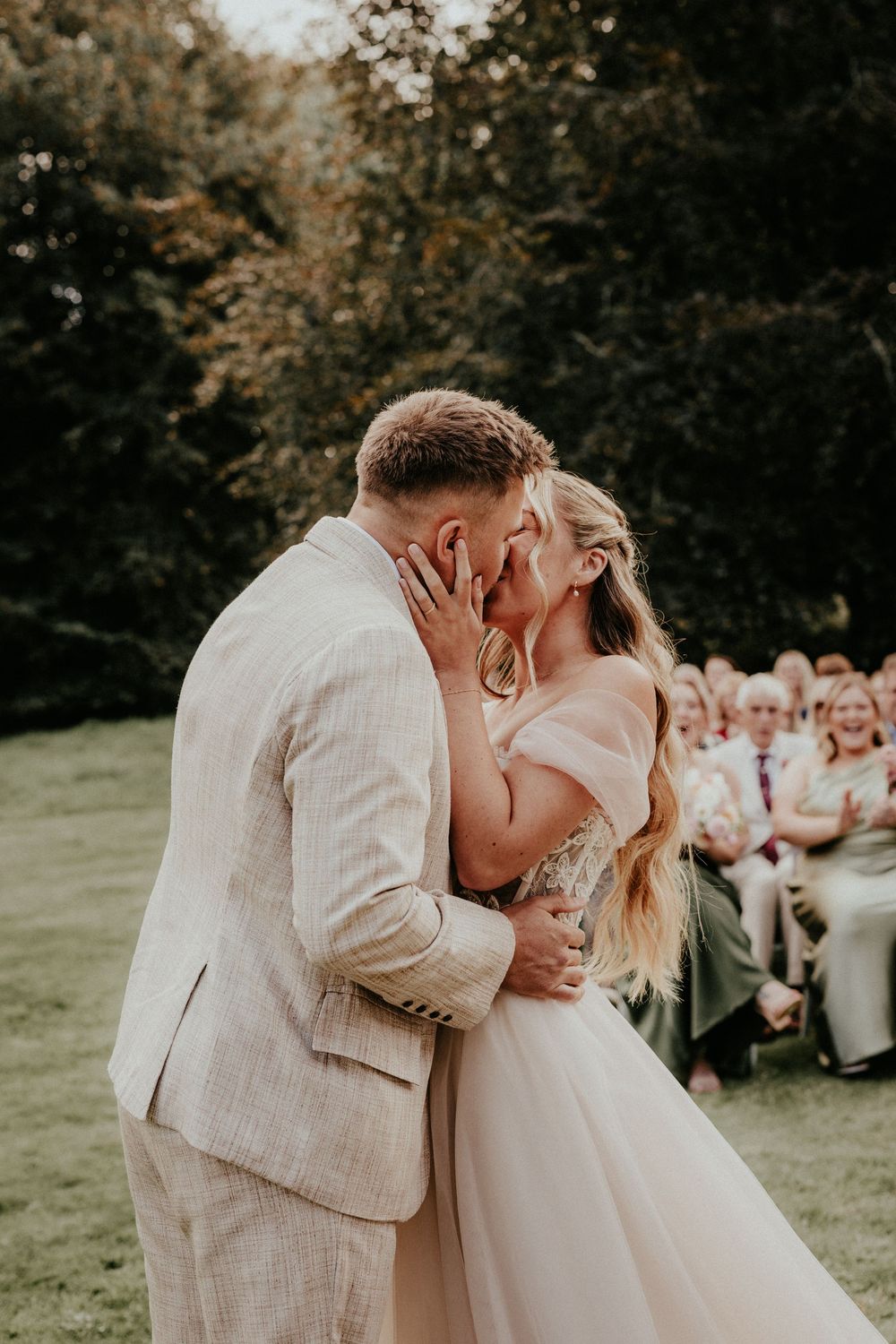 Newlyweds share a romantic kiss outdoors while wedding guests look on and celebrate in the background.