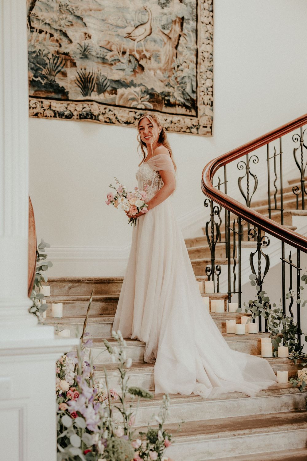 A bride in a flowing gown stands gracefully on an elegant white staircase adorned with floral decorations.