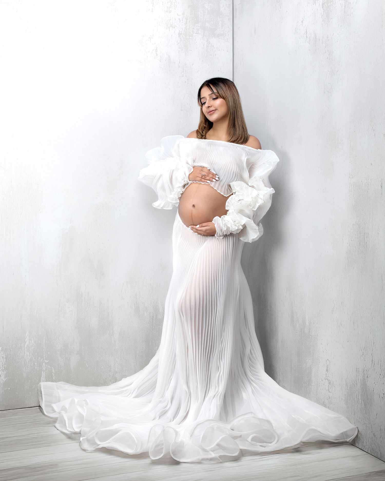 Woman in white skirt and top against a striking gray corner set holding her pregnant belly looking as gorgeous as ever.