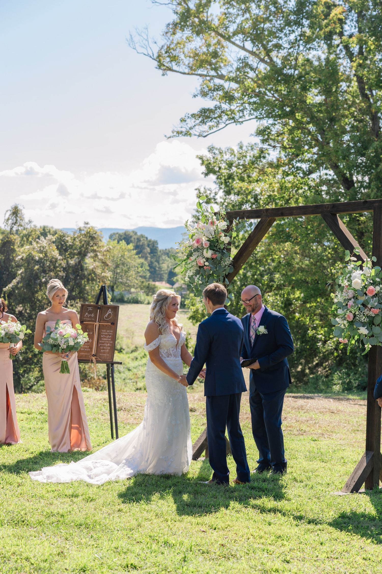 Wedding ceremony taking place outdoors on a sunny day with rolling hills and trees in the background.