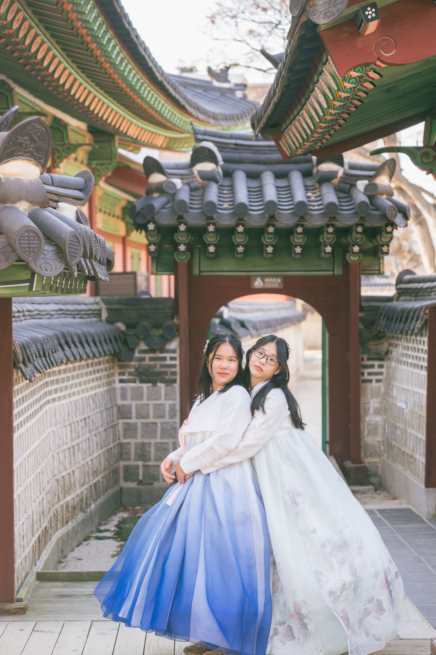 Traditional Korean hanbok dresses with blue ombre skirts displayed along a historic temple corridor with ornate architecture.