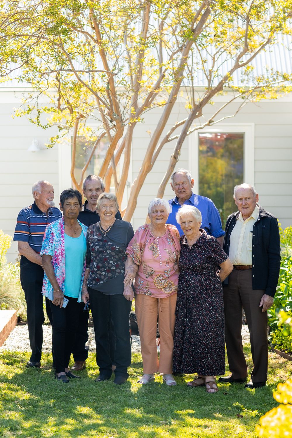A group of senior citizens standing together outdoors on a sunny day near a white building with autumn trees.