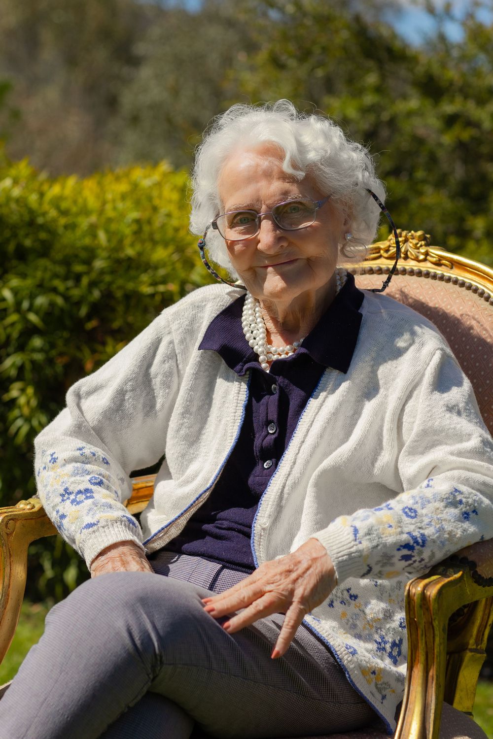 Elderly person in white cardigan and purple shirt resting on wooden garden bench in sunny outdoor setting.