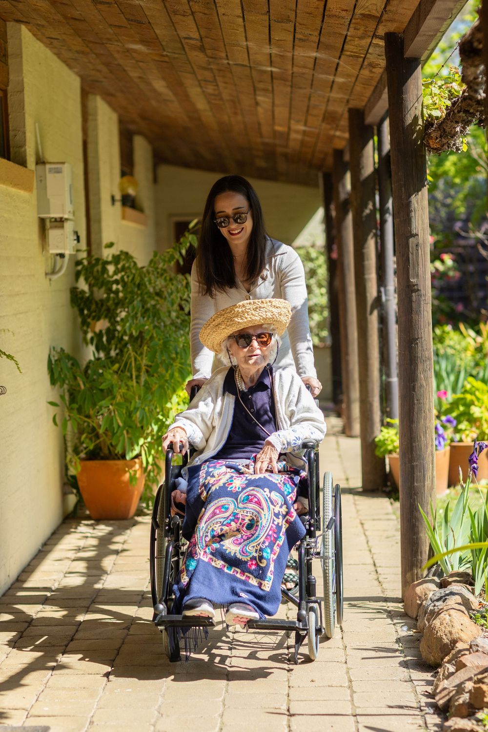 Person in wheelchair enjoys a peaceful moment under a covered outdoor walkway with wooden beams and potted plants.