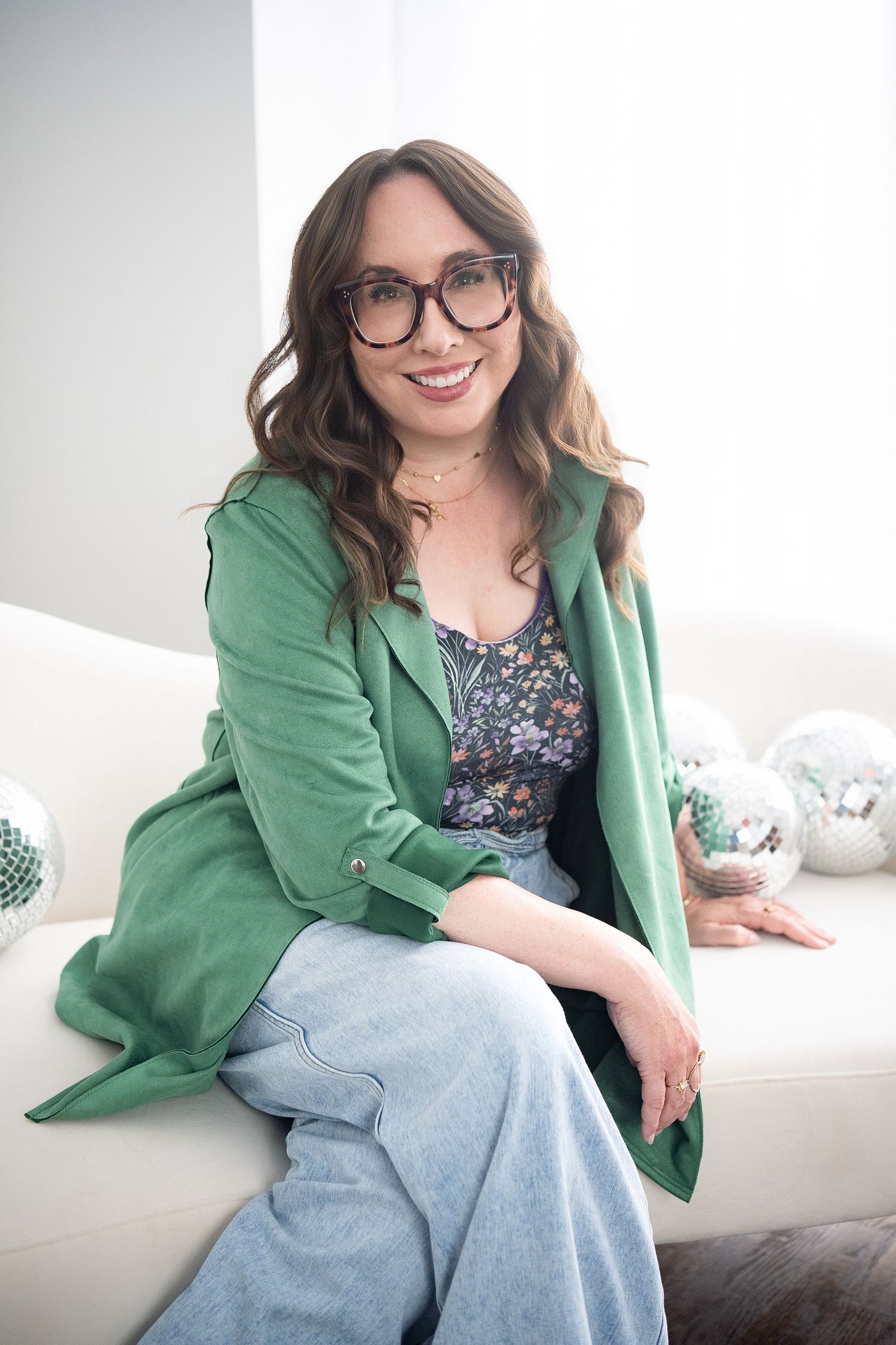 Woman in purple knit cardigan, floral top, and wide-leg jeans sitting on a cream sofa smiling.
