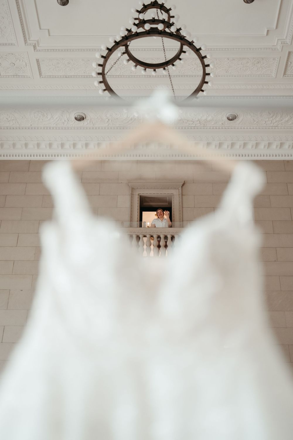 White wedding dress hanging on hanger with bride visible through window in elegant white venue.