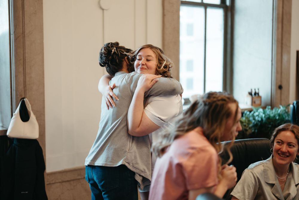 Two women sharing a warm hug in a bright indoor setting while others socialize nearby.