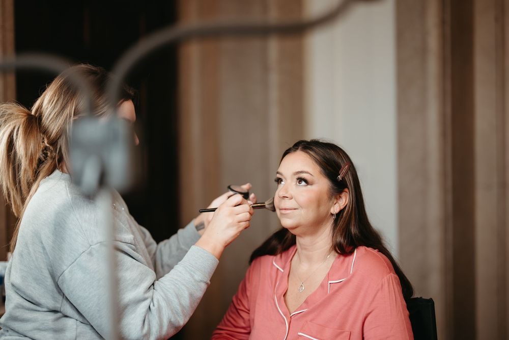 A makeup artist applies foundation to a woman in a pink pajama top, preparing her for a special occasion.