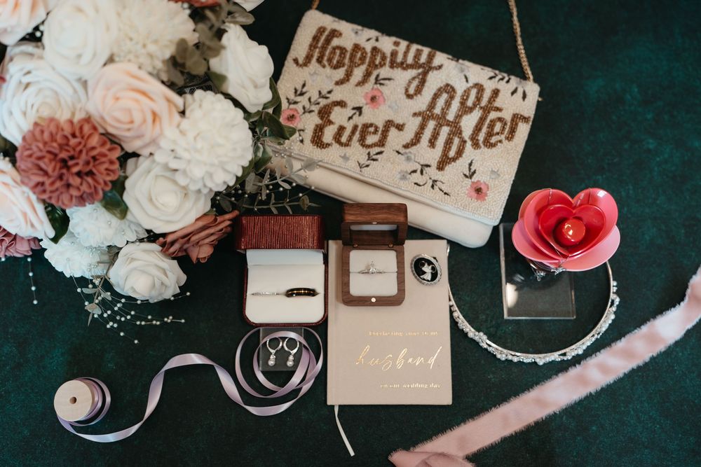 Wedding flat lay with floral bouquet, rings, wooden sign reading Happily Ever After, and elegant accessories on green velvet.