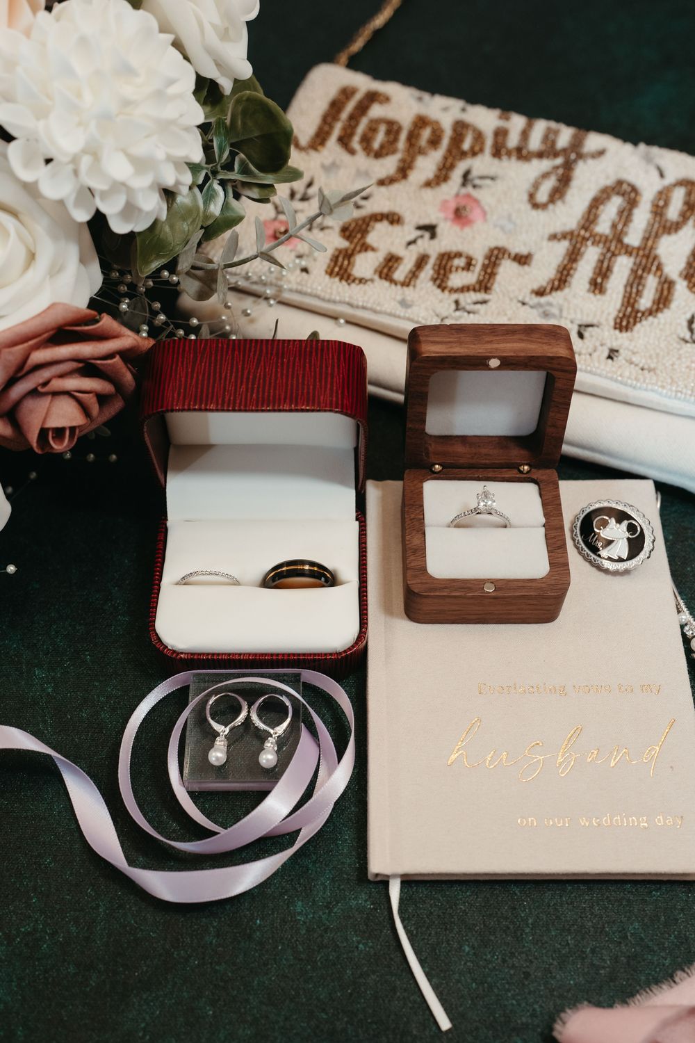 Wedding flat lay with diamond engagement ring in wooden box, gold bands, pearl earrings, and Happily Ever After clutch.
