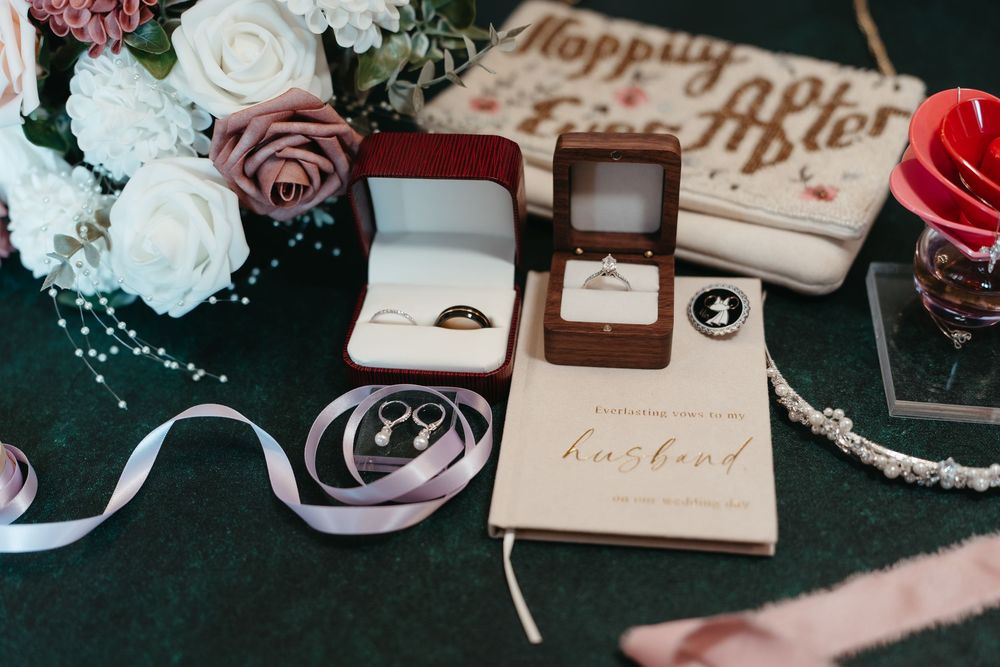 Wedding flat lay with ring boxes, gold bands, earrings, floral bouquet, ribbon, and a husband vow book on green velvet.