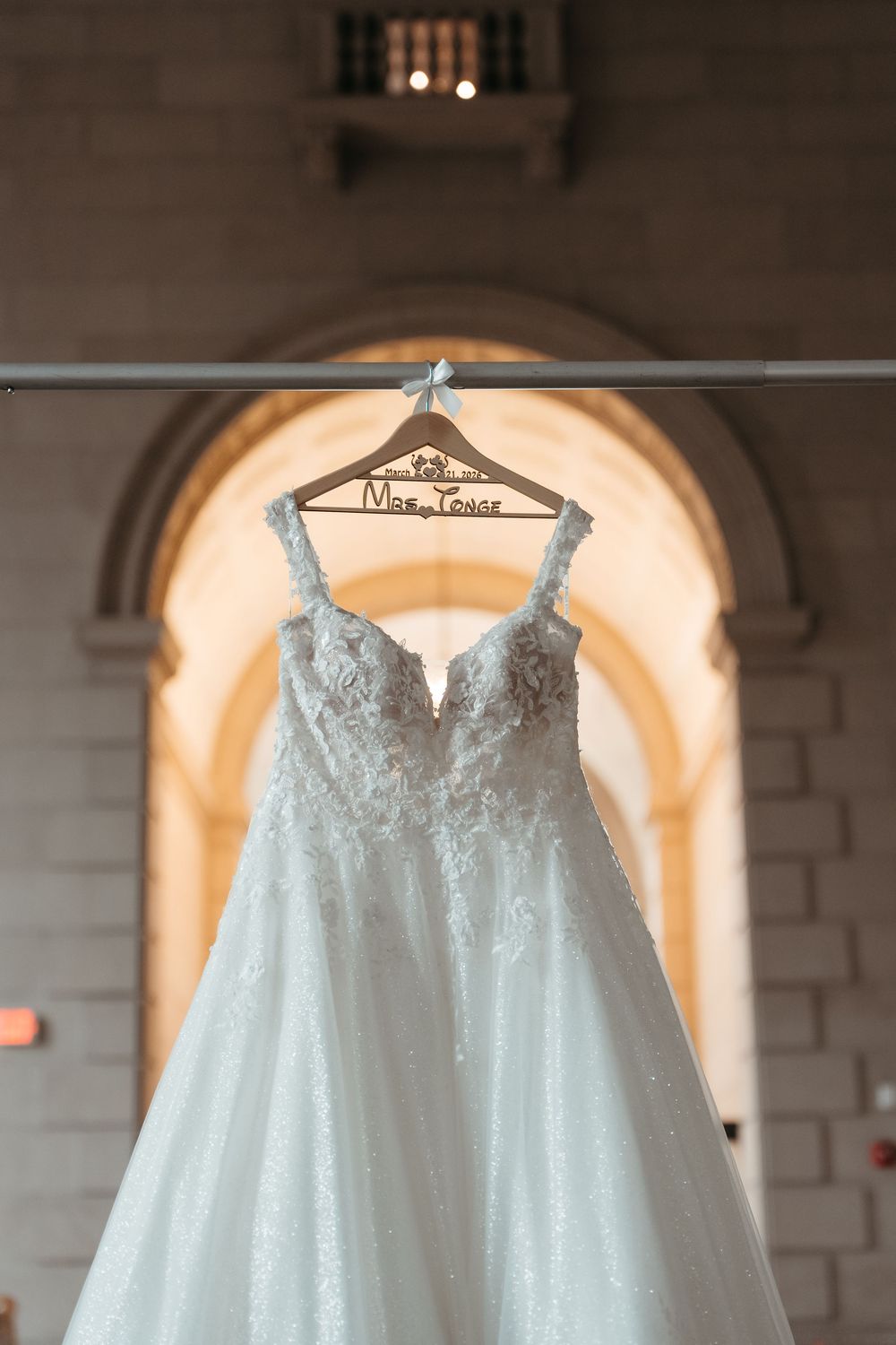 White lace wedding dress hanging on a personalized wooden hanger in an elegant arched venue.